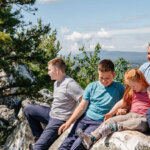 Holiday Inn Oak N Spruce 895×400 A family of five people sit on a rocky ledge overlooking a beautiful clear scenic view, after hiking.