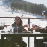 Winter skiing lunch at Bousquet credit Great Sky Media 895×400 A family of two adults and one child sit around a table eating lunch at the base of the snowy ski slopes of Bousquet Moutain.